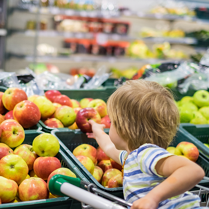 young c;aucasian boy reaches for a red apple in a grocery store as seen from behind.