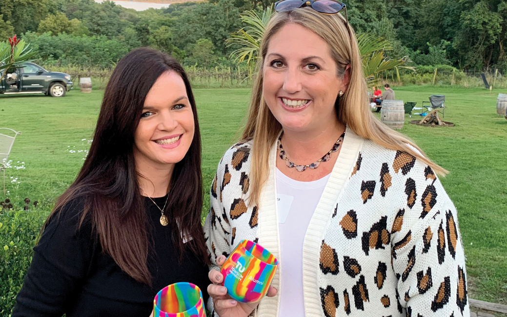 Two alumni standing outside, smiling and holding multicolored wine glasses