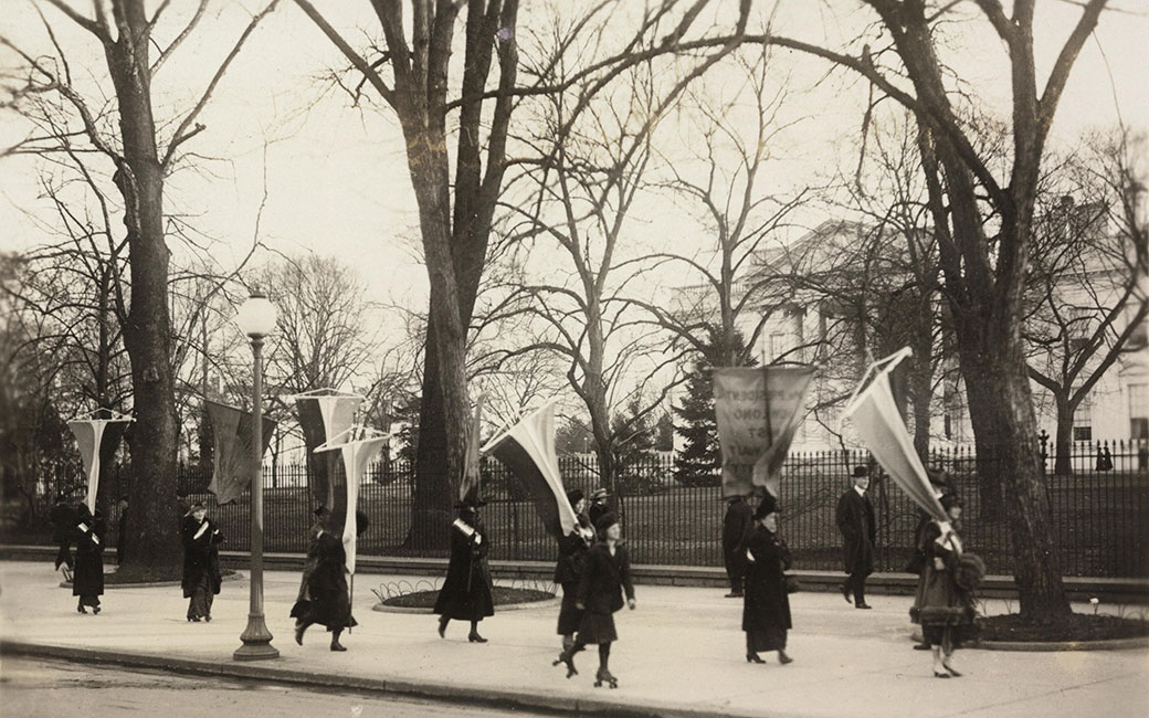 Black and white photo of suffragists with banners and flags on sidewalk outside of White House