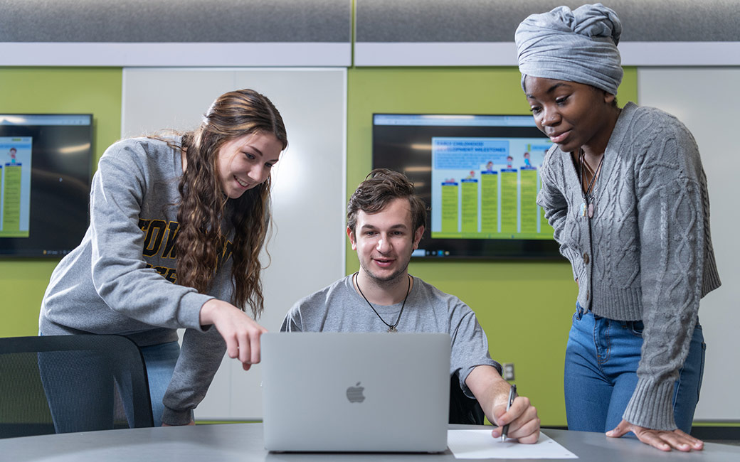 Three students point at laptop on table with monitor in background