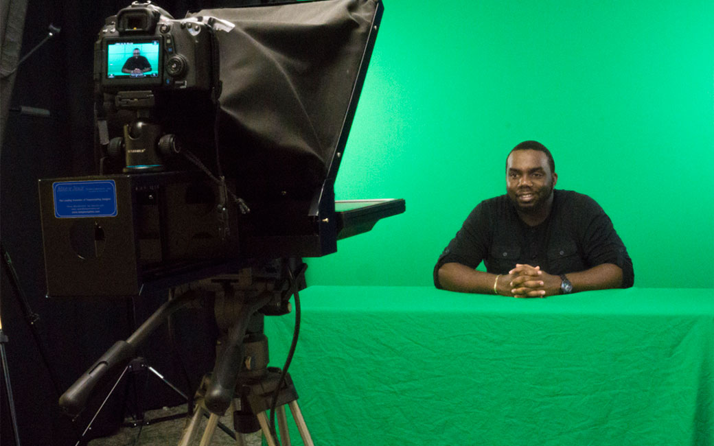 A student in Technology Support Desk studio C is seated in front of a teleprompter with a green screen behind 